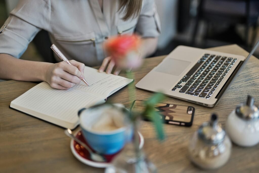 A writer taking notes in a paper notebook next to a laptop and a cup of coffee, illustrating an AI-powered creative workflow.