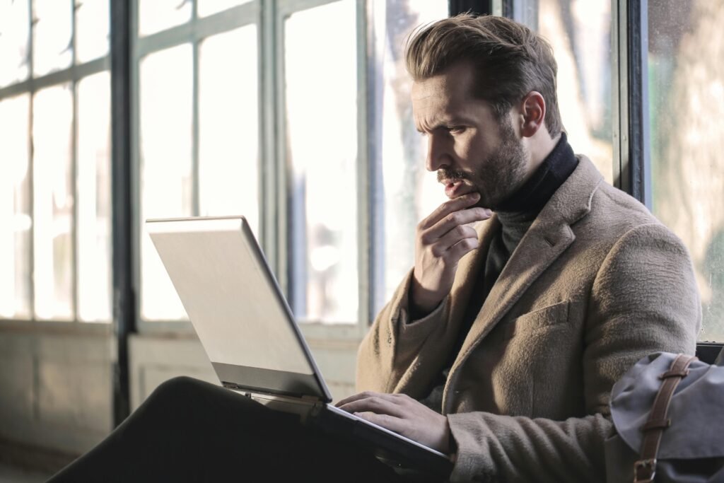 A fashionable man in a modern office looking at his laptop, weighing the pros and cons of using Grammarly for academic research.