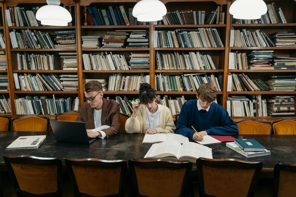 Three students engaged in studying together at a library table, focusing on books and laptops.