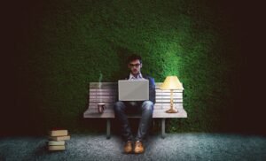 Scholar working on a laptop on a bench against a green hedge with a desk lamp and books.