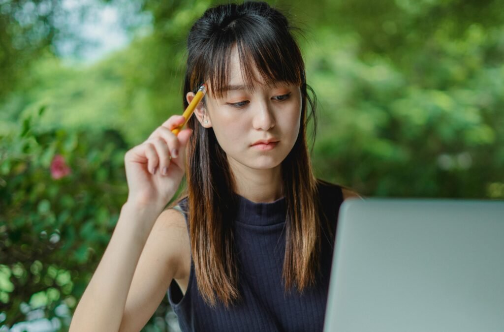 Asian student thinking carefully while working on laptop for academic writing.
