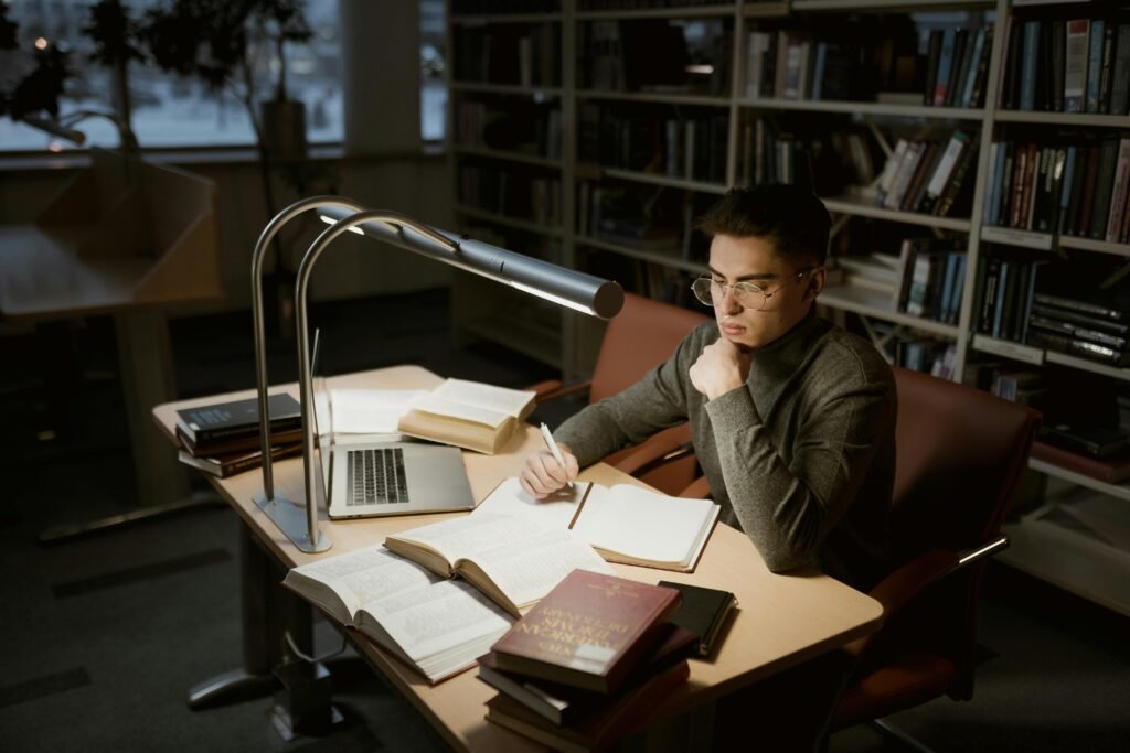 PhD student studying alone in dark library surrounded by books and laptop late at night.