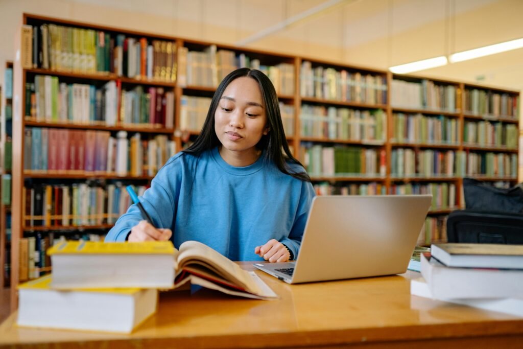 Student using Jenni AI on laptop in library with books for academic writing and research.