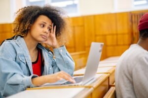 Student glancing sideways in lecture hall while working on laptop representing ambiguity around AI use and academic integrity.