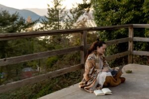 A researcher working on a laptop with an open book on a deck overlooking mountains and a body of water.