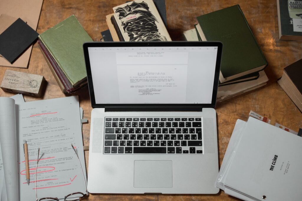 Top-down view of an open laptop surrounded by stacked books, reference papers, and annotated research drafts on a wooden desk.